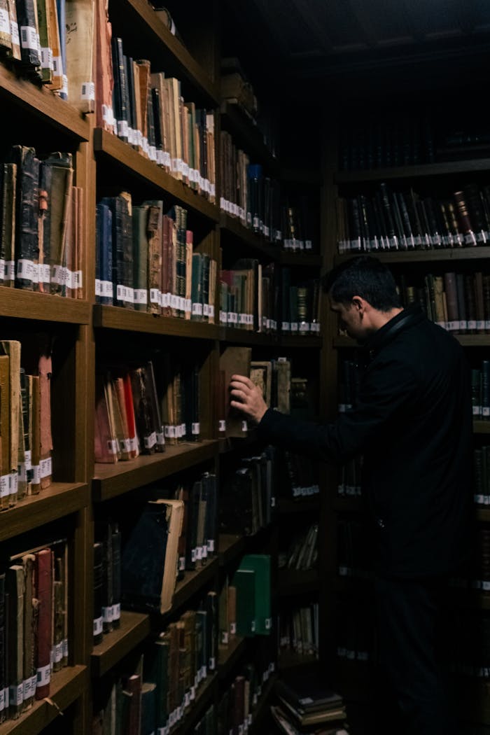 A man searching books in a dimly lit library aisle filled with old bookshelves.
