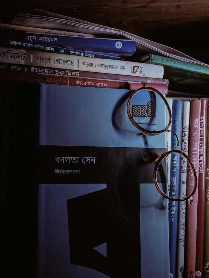 A dimly lit bookshelf with Bengali books and round glasses, evoking a vintage academic mood.